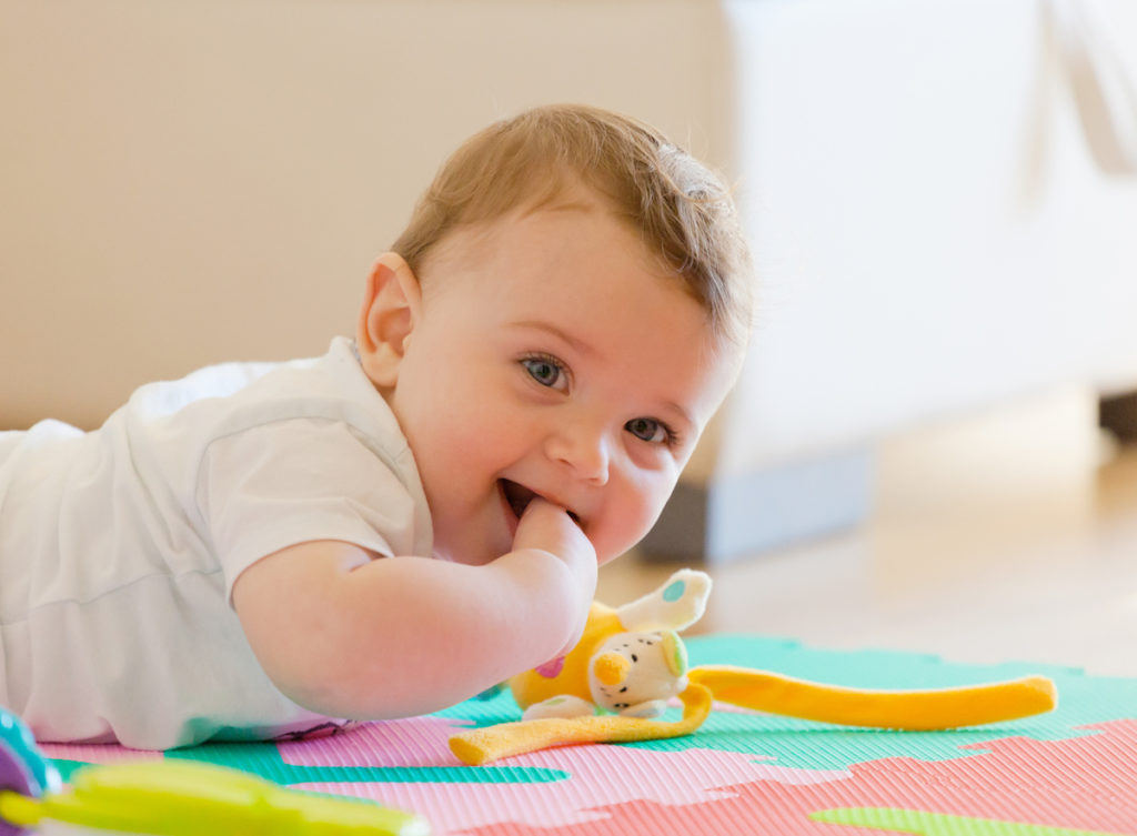 baby in a white onesie on his/her stomach on a play mat with a toy 